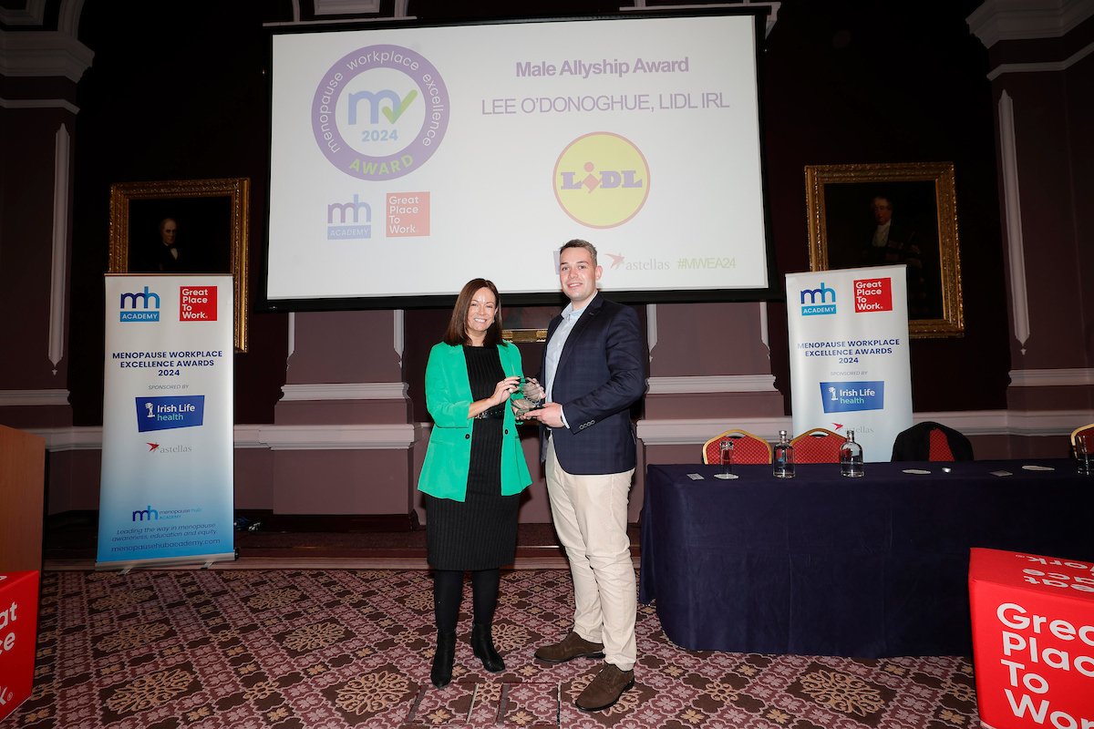 An award being presented on stage between two people, with Menopause Hub pull-up banners either side and a Male Allyship Award slide behind them