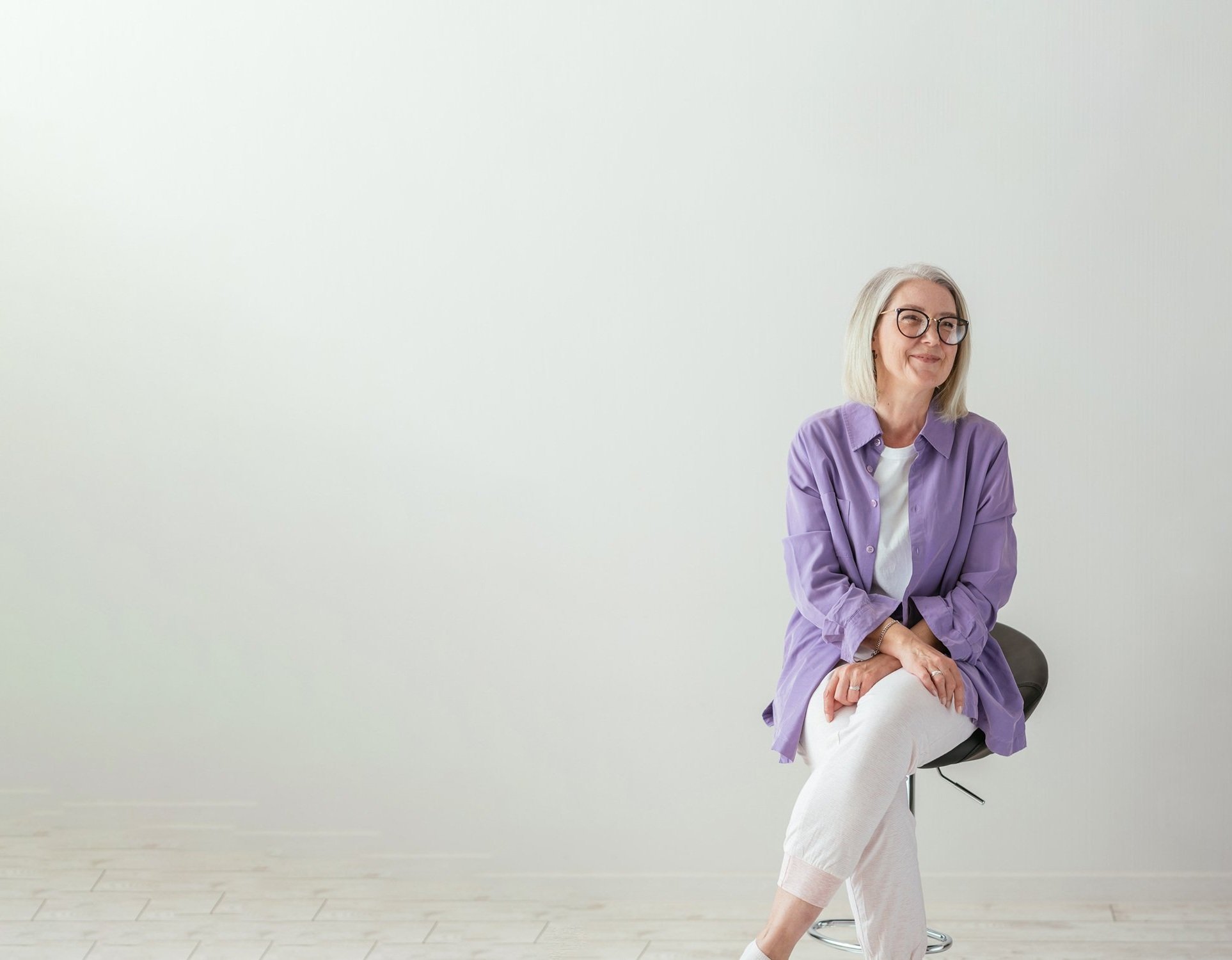 A woman with grey hair in a lilac shirt sitting on a stool against a white studio wall
