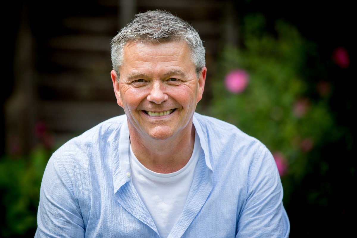 A smiling man with grey hair in a pale blue shirt photographed outdoors with greenery behind him